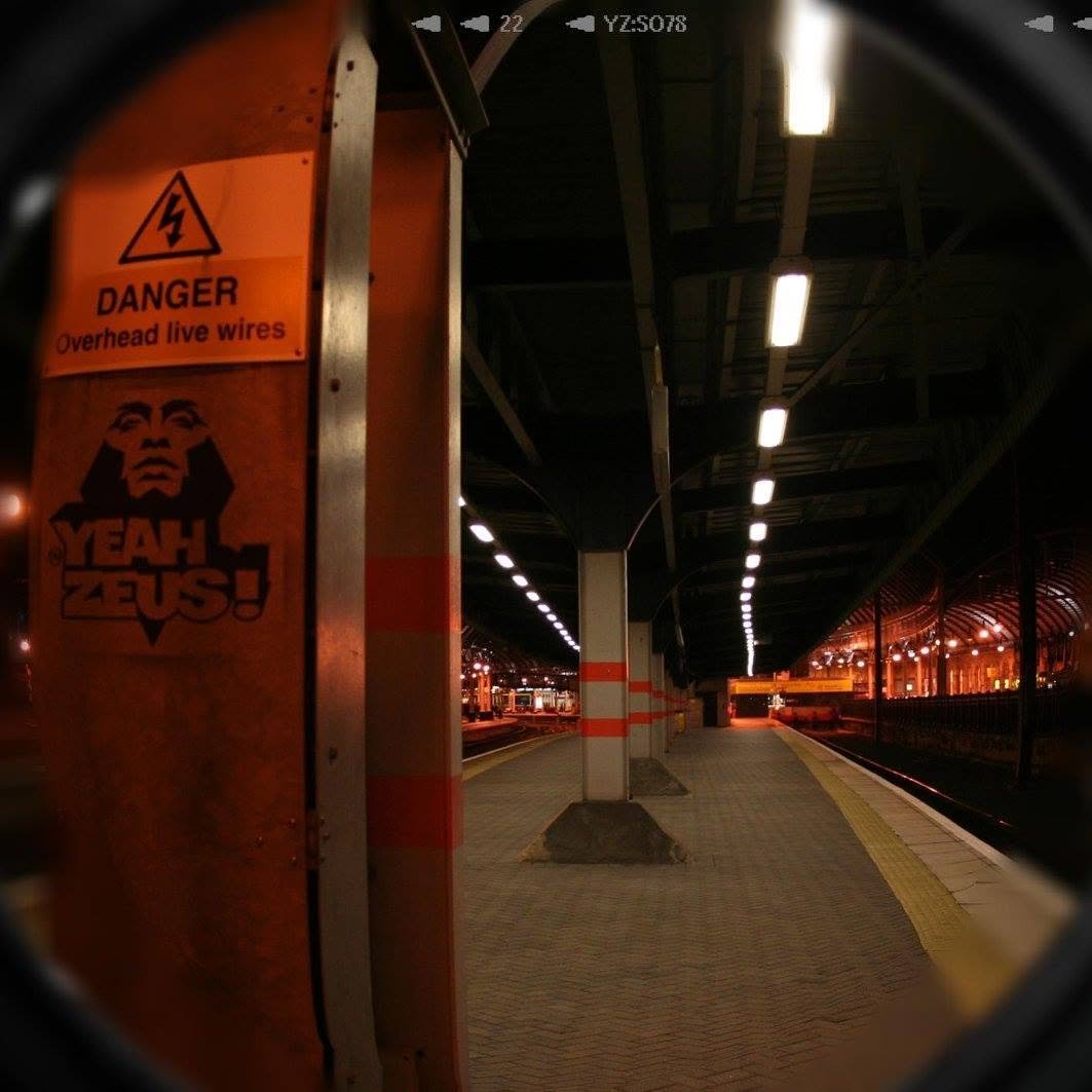 A late-night railway platform photographed through a fisheye lens, with a YEAH ZEUS stencil logo on the station wall.